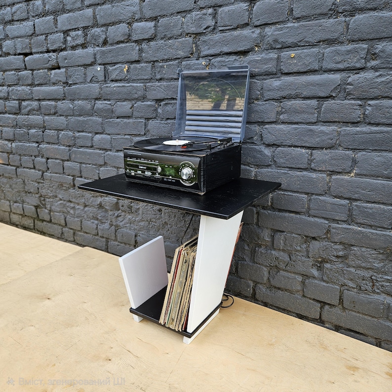 May include: A black and white record player stand featuring a flat top surface for a turntable and a lower angled storage compartment for vinyl records. The structure is constructed with a black top and base with a white angled support panel.