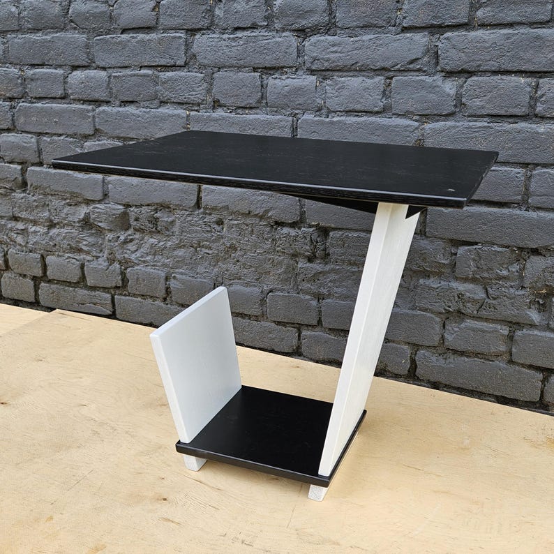 May include: A C-shaped side table featuring a rectangular black tabletop and a matching black base shelf. The structure is supported by a white angled leg and includes a white vertical panel attached to the base, creating a storage compartment.