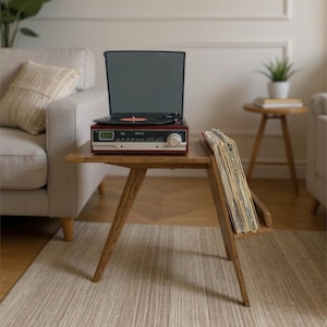 Könnte beinhalten: Ein Beistelltisch aus Holz mit schrägen Beinen und einem integrierten Ablagefach für eine Sammlung von Schallplatten. Ein dunkelroter und schwarzer Plattenspieler mit Digitalanzeige und einem großen silbernen Drehknopf steht auf der Tischplatte.