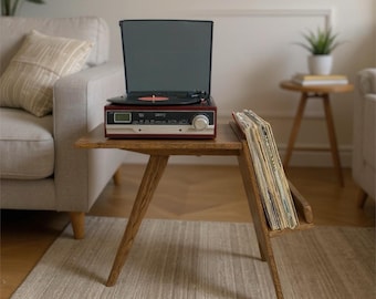 May include: A wooden side table with angled legs and an integrated side rack holding a collection of vinyl records. A maroon and silver record player with a black lid and a circular dial sits on the flat top surface of the wooden table.