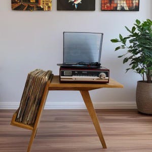 May include: A wooden record player stand with a built-in record player and a record holder. The record player is dark red with silver accents. A stack of vinyl records is in the holder. A potted plant is to the right.