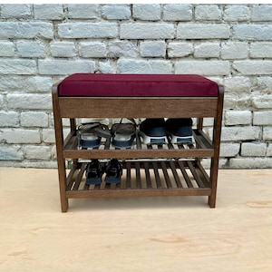 May include: A wooden shoe rack bench featuring a dark burgundy velvet upholstered seat cushion. The frame is constructed from brown wood with two slatted shelves for storage, currently holding four pairs of various shoes on the lower levels.