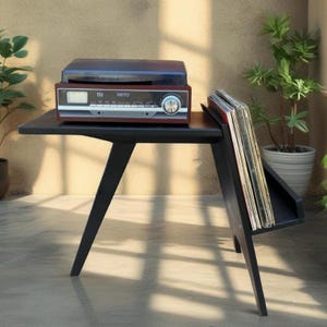 May include: A black wooden side table with angled legs and a built-in side rack holding several vinyl records. A dark wood and silver Camry brand record player with a clear plastic dust cover sits on the flat top surface of the table.