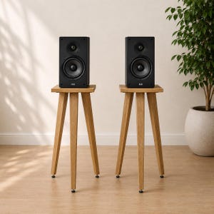 May include: Two black bookshelf speakers with the ELAC logo on the front, each mounted on a light-colored wooden tripod stand. The speakers feature a circular tweeter and a larger woofer driver, positioned on three-legged wooden bases.