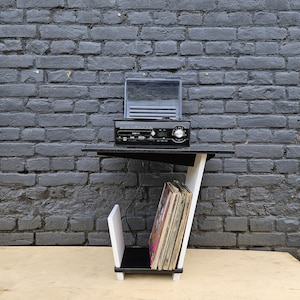 May include: A black and white side table featuring a top surface holding a black electronic record player with a clear lid. The lower section includes a vertical storage compartment containing a collection of vinyl records with visible album covers.