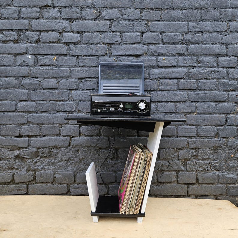 May include: A black and white side table featuring a top surface holding a black electronic record player with a clear lid. The lower section includes a vertical storage compartment containing a collection of vinyl records with visible album covers.