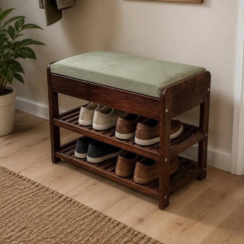 May include: A dark brown wooden shoe rack bench featuring a rectangular sage green upholstered seat cushion. The furniture piece includes two slatted shelves underneath, currently holding four pairs of casual sneakers in various neutral colors.