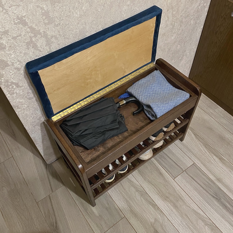 May include: A dark brown wooden shoe storage bench with a hinged lid featuring a blue velvet top. The interior compartment holds a black umbrella and a blue patterned fabric. A yellow wooden ruler is attached to the inner edge of the lid.