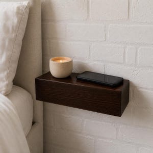 Op de afbeelding: Een rechthoekige donkerbruine houten zwevende plank gemonteerd aan een witte bakstenen muur. Een beige keramische kaarshouder met een brandende kaars en een zwarte smartphone staan op het vlakke oppervlak van de plank met zichtbare houtnerf.