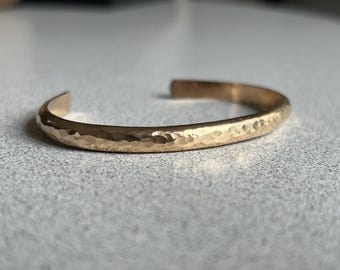 May include: A gold-toned metal cuff bracelet featuring a hammered, textured surface. The bracelet has an open-ended design with a rounded profile, showcasing a reflective finish that catches the light across its curved, metallic band.