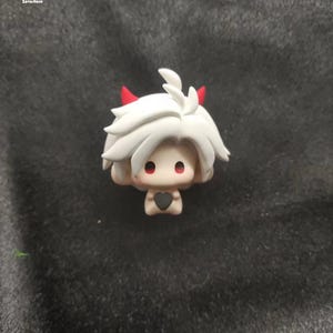 May include: A small white figurine with red horns and a black heart in its hands. The figurine has a white face with black eyes and a red mouth.