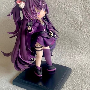 May include: Purple anime figurine with long hair, wearing a purple dress, black boots, and holding a gun. The figurine is standing on a black base.