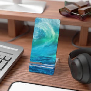 May include: A rectangular phone stand featuring a vibrant ocean wave design in shades of blue and green. The stand is made of a glossy material and is propped up by a silver-colored metal stand. The stand is on a wooden desk.