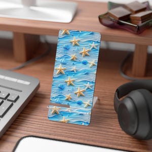 May include: A phone stand featuring a beach-themed design. The stand has a blue background with a wavy pattern, adorned with yellow starfish and blue seashells. The stand is made of a durable material and is designed to hold a phone upright.