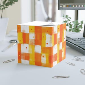 Könnte beinhalten: Ein quadratischer Notizblock mit einem farbenfrohen abstrakten Design. Das Muster zeigt orange, gelbe und weiße rechteckige Formen mit kleinen roten Punkten. Der Notizblock liegt auf einer weißen Oberfläche mit Bürobedarf.
