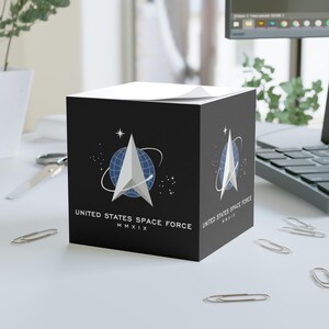 May include: A black cube-shaped notepad with the United States Space Force logo. The logo features a white arrow-shaped design over a blue globe, with the text "UNITED STATES SPACE FORCE MMXIX" in white.