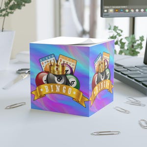 May include: A cube-shaped notepad with a colorful, swirling background. The design features bingo balls, cards, and a gold crown. A gold banner displays the word "BINGO".