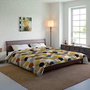 May include: A bed with a dark brown wooden frame and a colorful comforter. The comforter features an abstract pattern of large, irregular shapes in shades of yellow, brown, red, and black. White pillows and a painting are also visible.