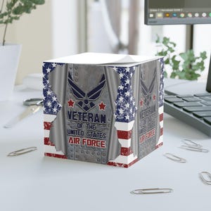 May include: A cube-shaped notepad featuring a design honoring U.S. Air Force veterans. The design incorporates the American flag's colors and stars, along with the Air Force emblem and text that reads "Veteran of the United States Air Force."