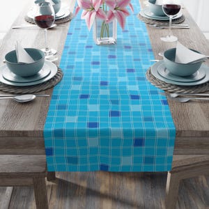 May include: A blue plaid table runner with a grid pattern, set on a wooden table. The runner features varying shades of blue and white lines. The table is set with plates, bowls, silverware, and wine glasses. A vase of pink flowers is in the center.