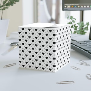 May include: A white cube-shaped notepad with a repeating pattern of black hearts and dots. The notepad is sitting on a white surface with paper clips scattered around it. A computer and plant are in the background.