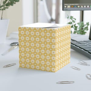 May include: A square notepad with a yellow and white star and checkerboard pattern. The notepad is on a white surface with paperclips and a computer in the background. The top of the notepad has a stack of white paper.