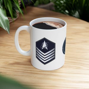 May include: White ceramic mug filled with dark coffee. The mug features a navy blue and silver emblem with a star and globe design, and three silver stripes. The mug is on a wooden surface.
