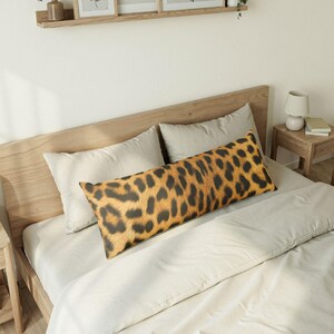 Puede incluir: Una almohada larga y rectangular con estampado de leopardo. La almohada presenta un fondo beige con manchas marrones oscuras, creando un diseño de estampado animal manchado. La almohada está colocada sobre una cama con ropa de cama de color neutro.