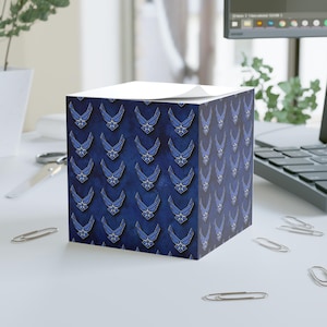 May include: A cube-shaped notepad with a repeating pattern of blue and silver Air Force emblems on a dark blue background. The notepad sits on a white surface with scattered paper clips.