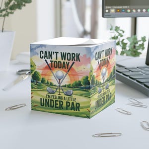 May include: A cube-shaped notepad with a golf-themed design. The artwork features crossed golf clubs, a golf ball, and the text "CAN'T WORK TODAY, I'M FEELING A LITTLE UNDER PAR" against a backdrop of a golf course at sunset.