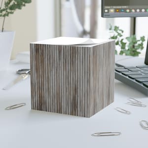 Puede incluir: Un bloc de notas cúbico con textura de vetas de madera. El bloc de notas tiene una pila de papel blanco en la parte superior. Varios clips plateados están esparcidos alrededor del bloc de notas sobre una superficie blanca. Un ordenador y unas tijeras están en el fondo.