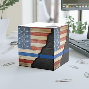 May include: A cube-shaped notepad featuring a distressed American flag design. The design includes red and white stripes, blue stars, and a blue line. The notepad is on a white surface with office supplies.