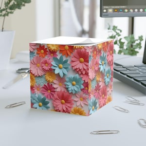 May include: A floral-themed cube-shaped notepad featuring a vibrant array of colorful paper flowers. The flowers are in shades of pink, blue, orange, and yellow, creating a cheerful and decorative design. The notepad sits on a white surface.