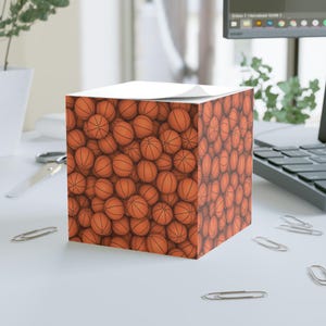 May include: A cube-shaped notepad with a repeating pattern of orange basketballs. The notepad has a white top sheet and is surrounded by office supplies on a white desk.