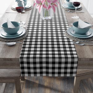 May include: A black and white checkered table runner on a wooden table. The runner is rectangular and covers the center of the table. The table is set with plates, bowls, silverware, and wine glasses. A vase of pink flowers sits in the center.