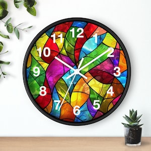 Puede incluir: Un reloj de pared redondo con marco negro. La esfera del reloj presenta un diseño de vidriera de colores con tonos de rojo, amarillo, verde, azul y morado. Los números y las manecillas blancas indican la hora. Una pequeña planta en maceta está sobre una superficie de madera.