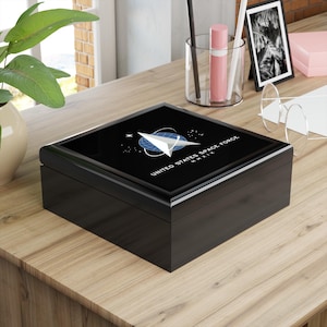 May include: A black wooden box with a glossy finish. The lid features the United States Space Force logo in white and blue against a black background. The text "UNITED STATES SPACE FORCE MMXIX" is printed on the lid.