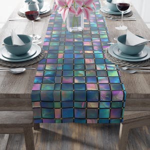 May include: A table runner with a mosaic tile design in shades of blue, purple, and gold. The runner is placed on a wooden table set for a meal, with plates, glasses, and flowers.