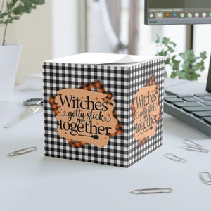 May include: A cube-shaped notepad with a black and white checkered pattern. The notepad features the text "Witches gotta stick together" in a stylized font, with orange and brown accents. A witch's broom graphic is also included.