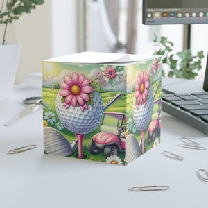 May include: A decorative cube featuring a golf theme. The design includes a golf ball with a pink flower, a pink golf cart, and golf clubs on a green golf course. The cube is adorned with daisies and other flowers.