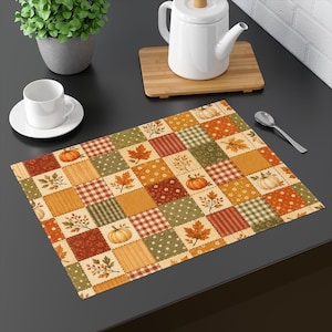 May include: A rectangular placemat featuring a patchwork design of autumn-themed squares. The pattern includes orange pumpkins, brown maple leaves, green foliage, and various plaid, striped, and polka dot textures in shades of orange, brown, and green.