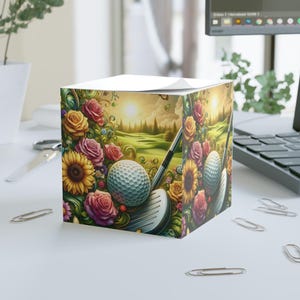 May include: A golf-themed sticky note cube featuring a golf ball, club, and a golf course scene with roses and sunflowers. The cube is decorated with a vibrant floral design and a golf course landscape.