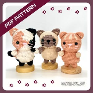 Op de afbeelding: Drie gehaakte amigurumi-katten speelgoed, elk met een ander kleurenschema. De katten staan op houten bases. De afbeelding bevat de tekst "COFFEE.CO.ART - ARTISAN CROCHET - ".