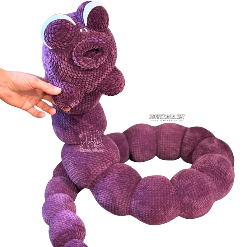 May include: A handmade, plush toy in the shape of a purple worm. The toy has a large head with white-rimmed eyes and a wide mouth. The segmented body is a deep purple colour. The toy is designed to be soft and cuddly.