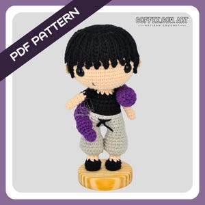 May include: A handmade crochet doll with black hair, a black shirt, and gray pants. The doll has purple accents and is standing on a wooden base. The image includes the text "PDF PATTERN" and "COFFEE.COM. ART - ARTISAN CROCHET".