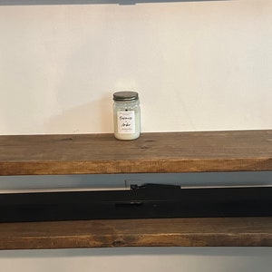 Puede incluir: Una estantería de madera rústica con una barra de sonido negra, un tarro de vela blanca y un libro titulado "Minimalist" en la repisa superior. La estantería tiene un hueco en el medio para la barra de sonido.