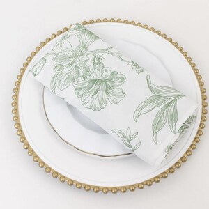 May include: A white plate with a gold beaded rim and a smaller white plate inside. A folded white napkin with a green floral design rests on the plates. The napkin features a botanical illustration of flowers and leaves.
