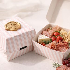 May include: A charcuterie box with pink and white striped packaging. The box contains an assortment of meats, cheeses, crackers, and fresh strawberries. A gold-colored wax seal adorns the top of the closed box.