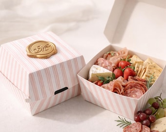 May include: A charcuterie box with pink and white striped packaging. The box contains an assortment of meats, cheeses, crackers, and fresh strawberries. A gold-coloured wax seal adorns the top of the closed box.