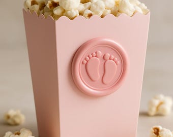 May include: A pink popcorn box filled with popcorn, featuring a baby footprint design. The box has a scalloped top edge and a circular emblem with two baby footprints. Scattered popcorn pieces are around the box.